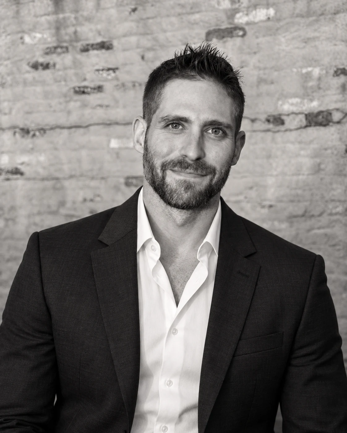 Black-and-white portrait of Brad Dworkin in a dark suit jacket and open white shirt, seated against a brick wall.